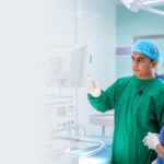 A RAGEC surgeon in a blue surgical cap and green gown reviewing laparoscopic equipment with a masked surgical team in a modern operating room.
