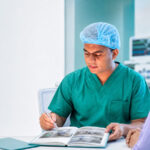 A gynecologist naturally holding a pen and open medical book while explaining hospital facilities and laparoscopic care to a woman in a bright consultation room.