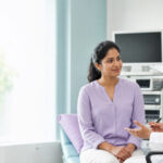 A patient speaking with a gynecologist in a bright consultation room with laparoscopic equipment visible in the background.